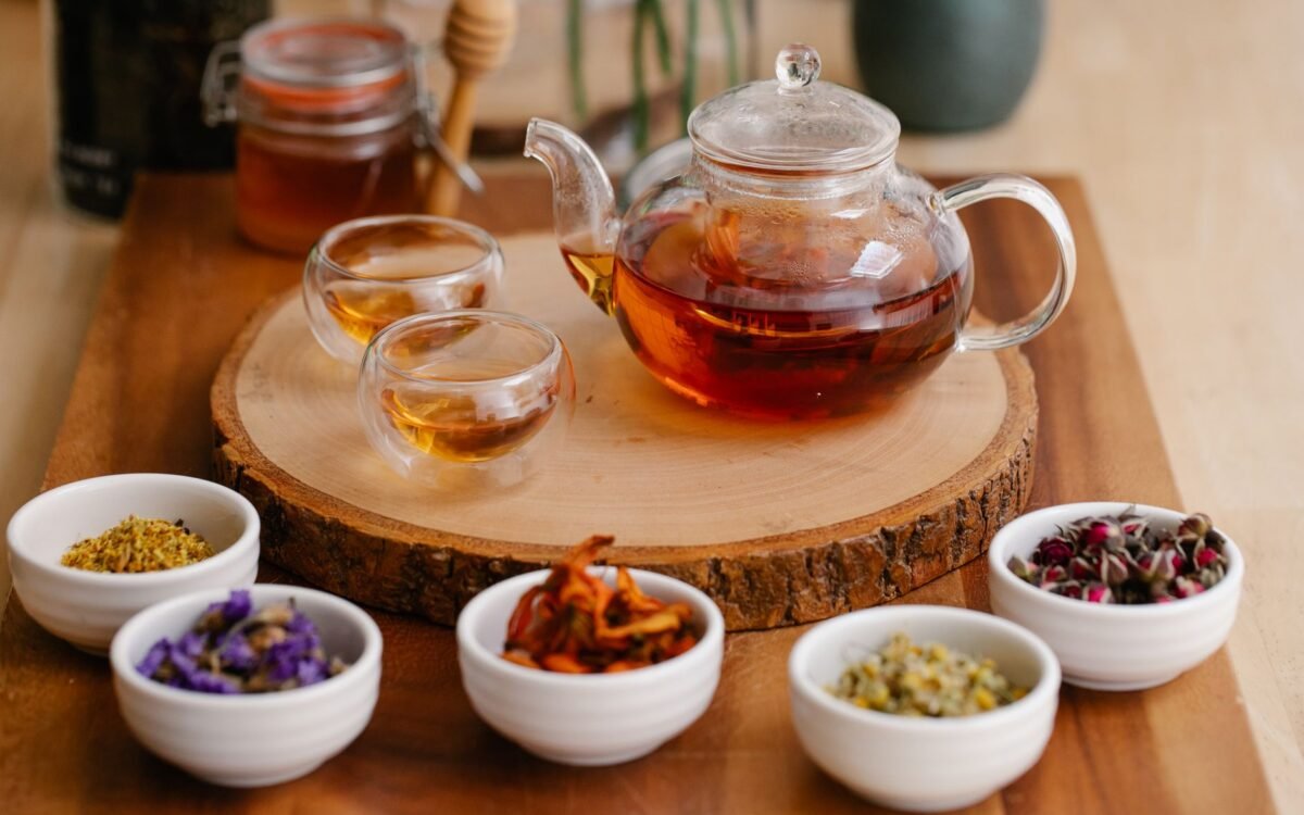 Bule de vidro e xícaras com infusões de ervas, ervas frescas e colheres de mel em uma bandeja de madeira
