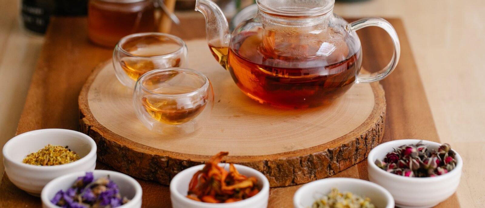 Bule de vidro e xícaras com infusões de ervas, ervas frescas e colheres de mel em uma bandeja de madeira