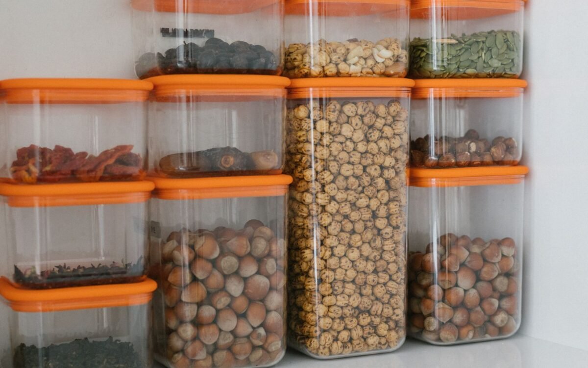Diversos grãos e castanhas armazenados em recipientes de plástico transparente com tampas laranja, organizados ordenadamente