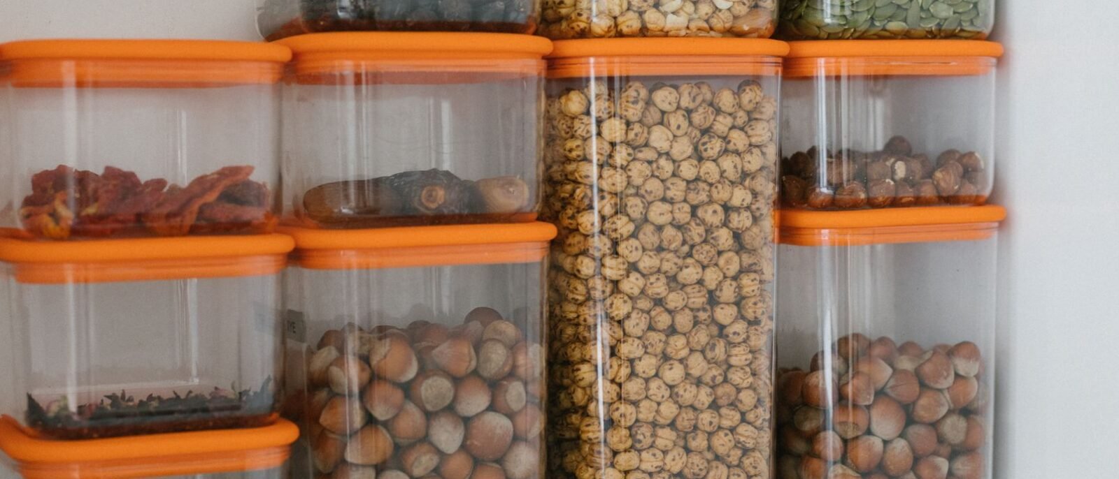 Diversos grãos e castanhas armazenados em recipientes de plástico transparente com tampas laranja, organizados ordenadamente